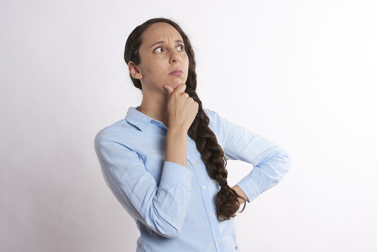 A woman with brown hair in a braid wearing a light blue button up shirt with a fist on her hip and the other hand on her chin thinking
