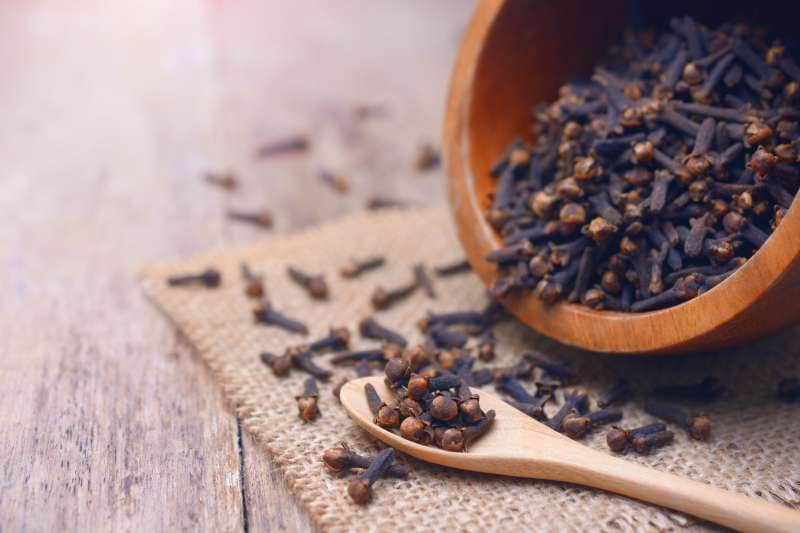 Cloves pouring out of a wooden bowl onto a wooden spoon on a wood table.